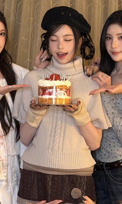Three young women pose together indoors, with the central figure wearing a black beret and cream turtleneck while holding a small cake topped with strawberries and lit candles, flanked by two friends who frame her with their hands. The caption about "mcm c Ian did a plum hairstyle" reflects the braided pigtails styled into the central woman's hair, giving a soft, cute aesthetic to the celebratory moment.