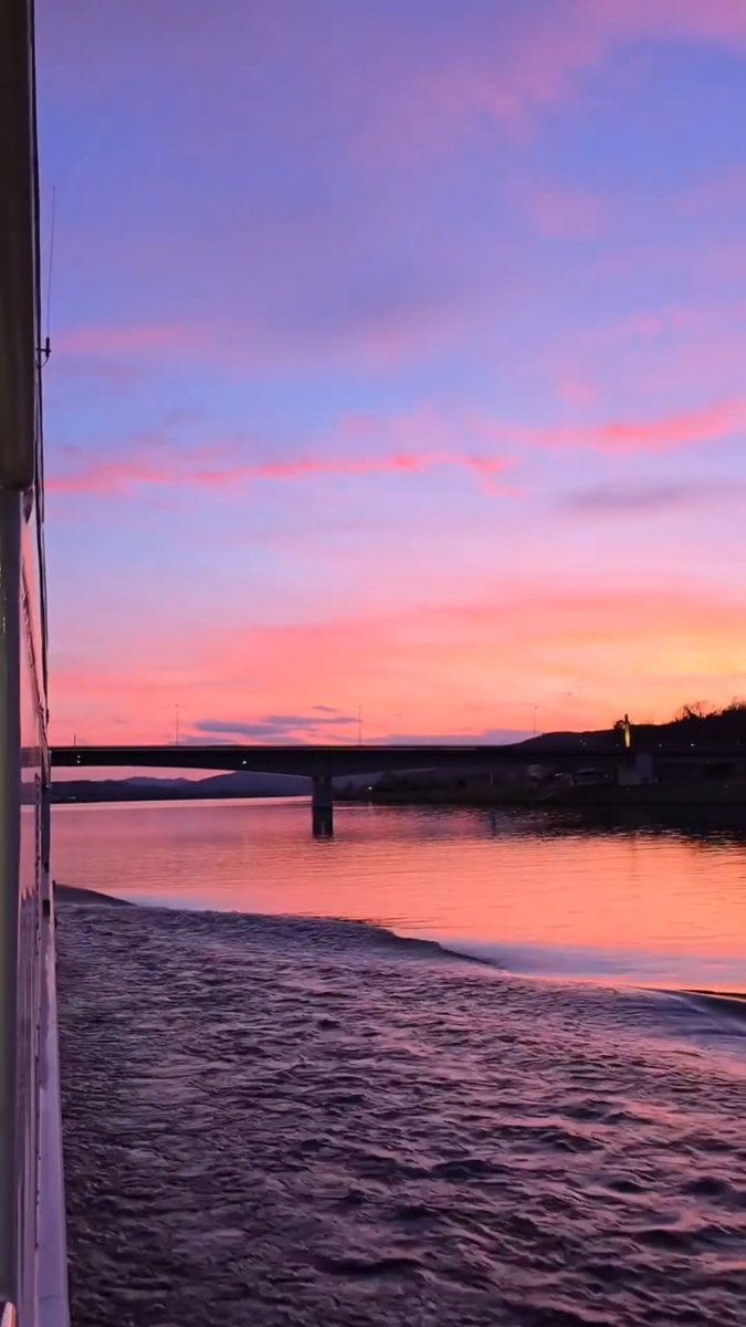 A vertical photo of a pink and purple sunset reflecting on a body of water, with a bridge visible in the distance and gentle waves in the foreground.