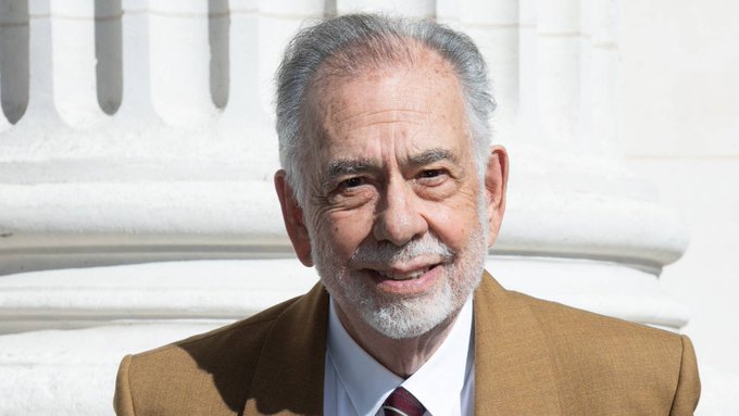 An elderly man with a white beard wearing a tan suit and striped tie smiles warmly in front of white stone columns.