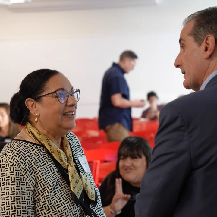 A person wearing glasses and patterned clothing is speaking with another person in a suit. They are in a room with red chairs and several people sit in the background. The atmosphere appears conversational and informal.