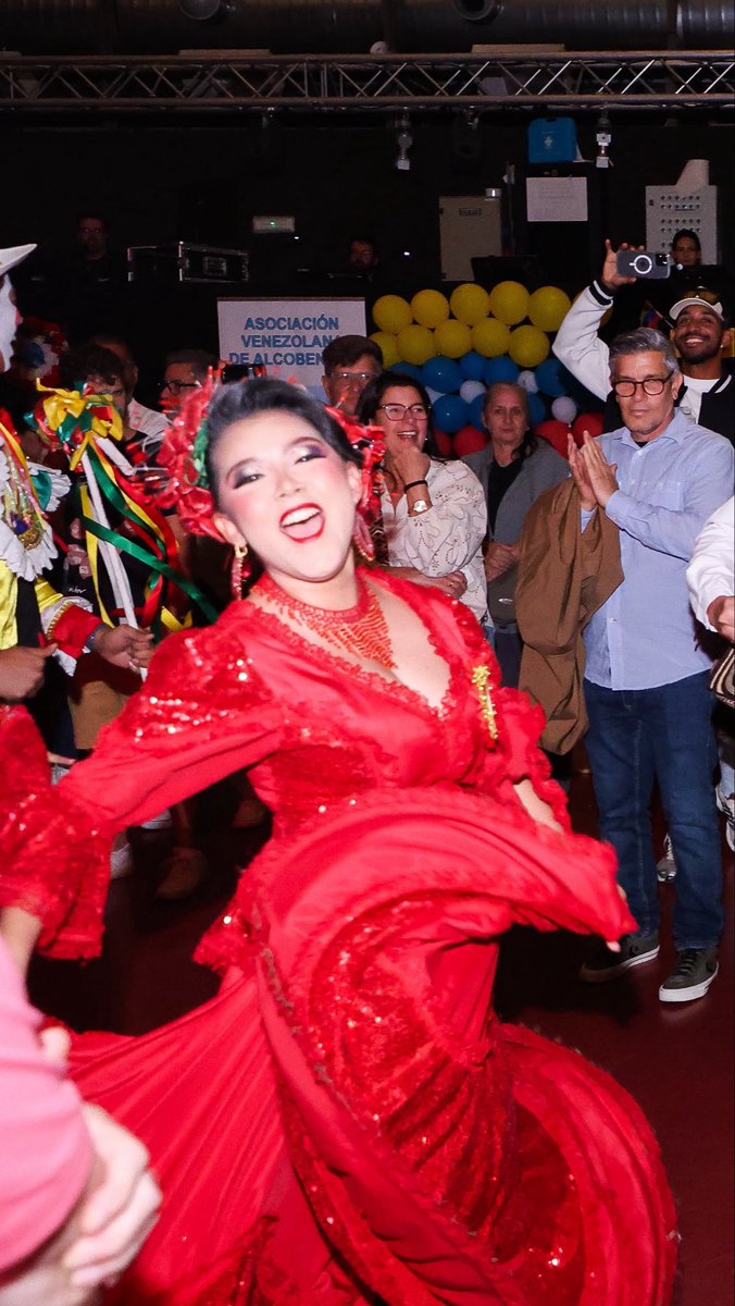 A woman in a vibrant red traditional dress with ribbons in her hair dances at a Venezuelan association event, with a banner reading "ASOCIACIÓN VENEZOLANA DE ALCOBEN" visible behind her.
