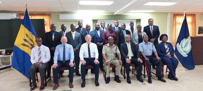 A formal group photograph features approximately twenty people in business attire and uniforms posed in an office setting between the Barbados flag and another official flag.
