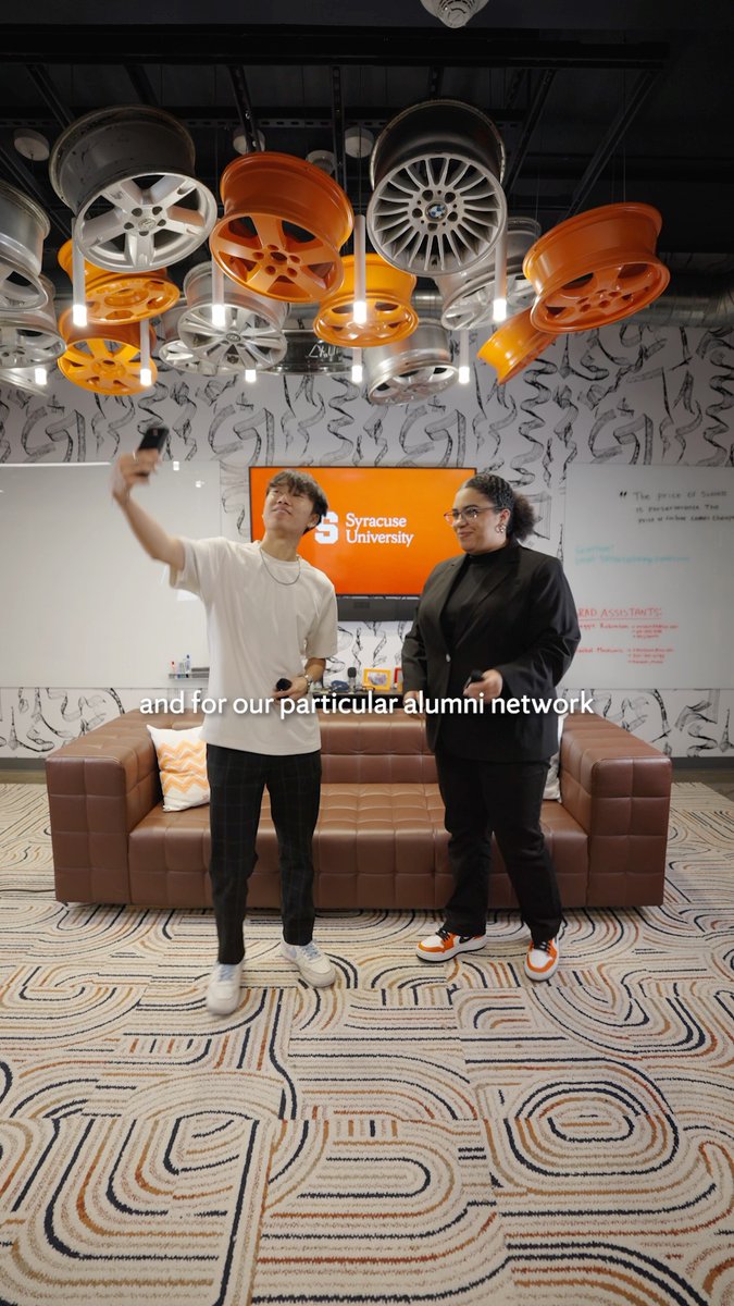 Two people standing in a lounge area with car wheel hubcaps hanging from the ceiling, one holding up a phone for a selfie, with a 'Syracuse University' TV screen behind them and text overlay 'and for our particular alumni network'.
