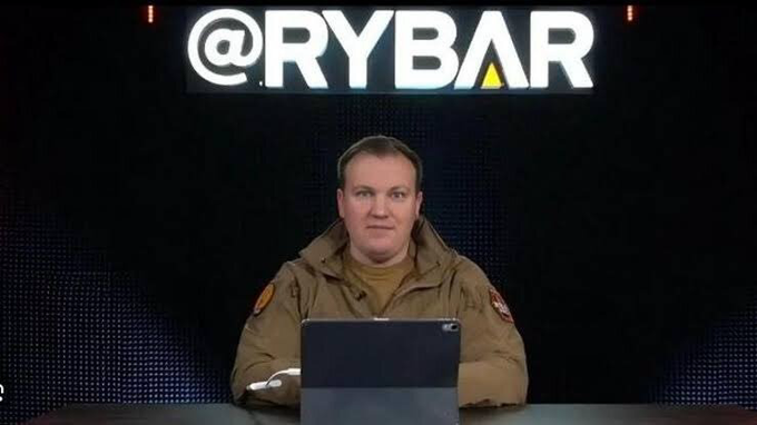 A man in a tan military-style jacket sits at a desk with a tablet in front of a large illuminated "@RYBAR" sign, representing Mikhail Zvinchuk, the founder of the pro-Kremlin media network Rybar, who was detained in Montenegro.