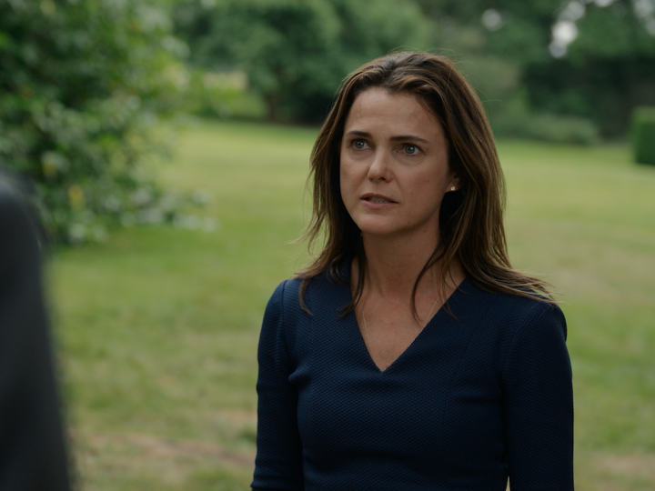 A woman with brown hair wearing a navy V-neck sweater stands outdoors in a grassy area, appearing to be engaged in a serious conversation with someone off-camera. This appears to be a scene from the Netflix series "The Diplomat," showcasing one of the show's dramatic outdoor moments as part of a countdown to Season 4.