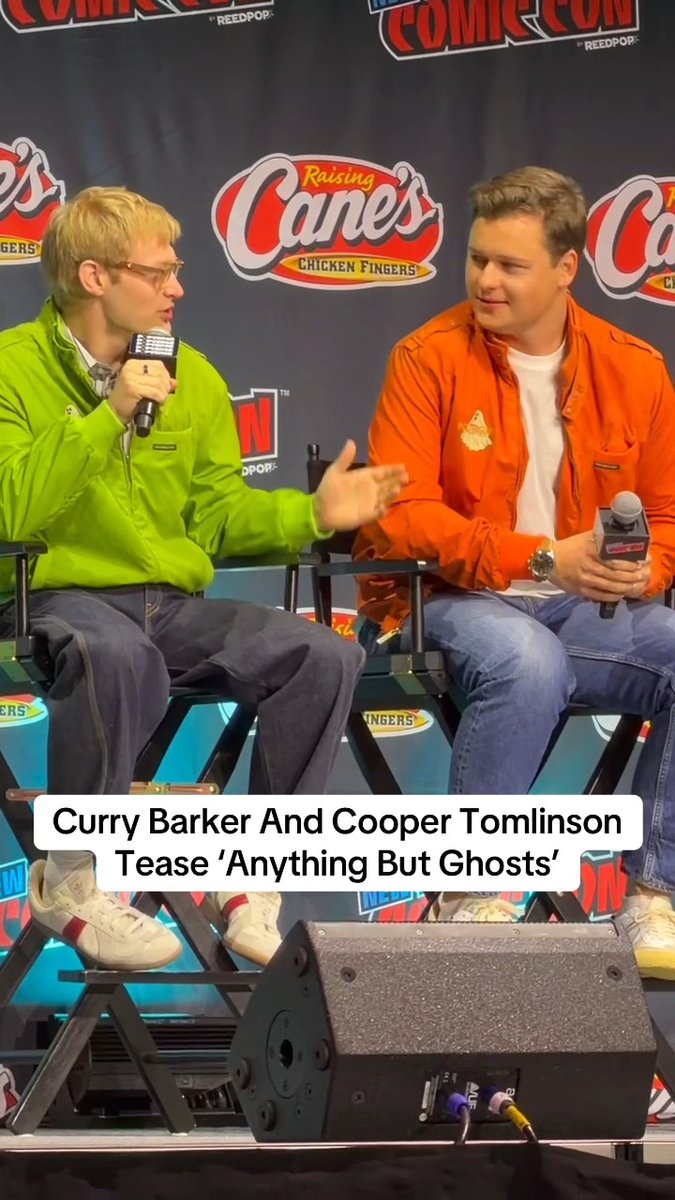 Two men sit on a panel at a Comic Con event with Raising Cane's sponsorship banners behind them, with the caption "Curry Barker And Cooper Tomlinson Tease 'Anything But Ghosts'" displayed. One man wears a green jacket while the other wears an orange jacket, both holding microphones as they discuss the film.