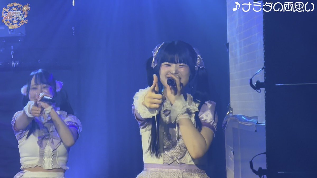 Two performers in white and purple idol costumes sing on a blue-lit stage, with one in the center holding a microphone and pointing at the camera while smiling; Japanese text '♪きざしの雨の中で' appears in the top right corner.