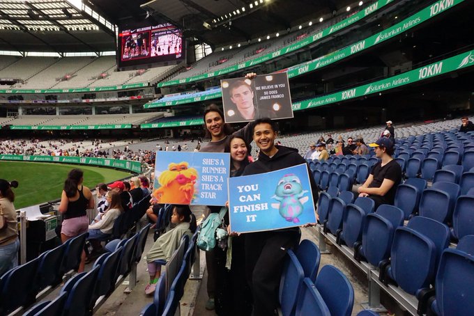 Fans in a stadium holding up signs reading 'WE BELIEVE IN YOU', 'SHE'S A RUNNER SHE'S A TRACK STAR', and 'YOU CAN FINISH IT' alongside photos, at a Sydney event.