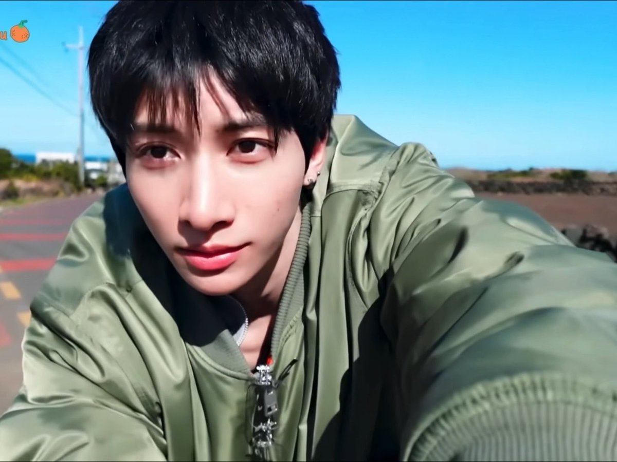 A young man with dark hair wearing an olive green puffy jacket takes a selfie outdoors, with a small orange logo in the upper left corner.