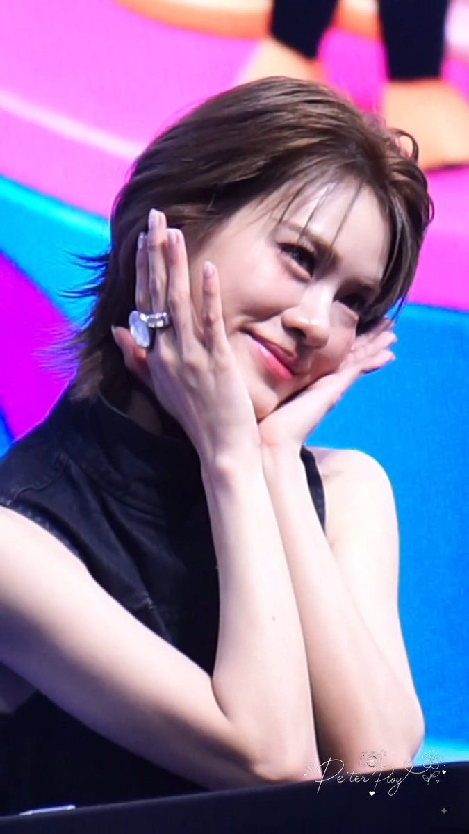 A young woman with short brown hair poses playfully with her hands framing her face, wearing a black sleeveless top and a large ring, with a watermark 'Peter Huy' in the corner.