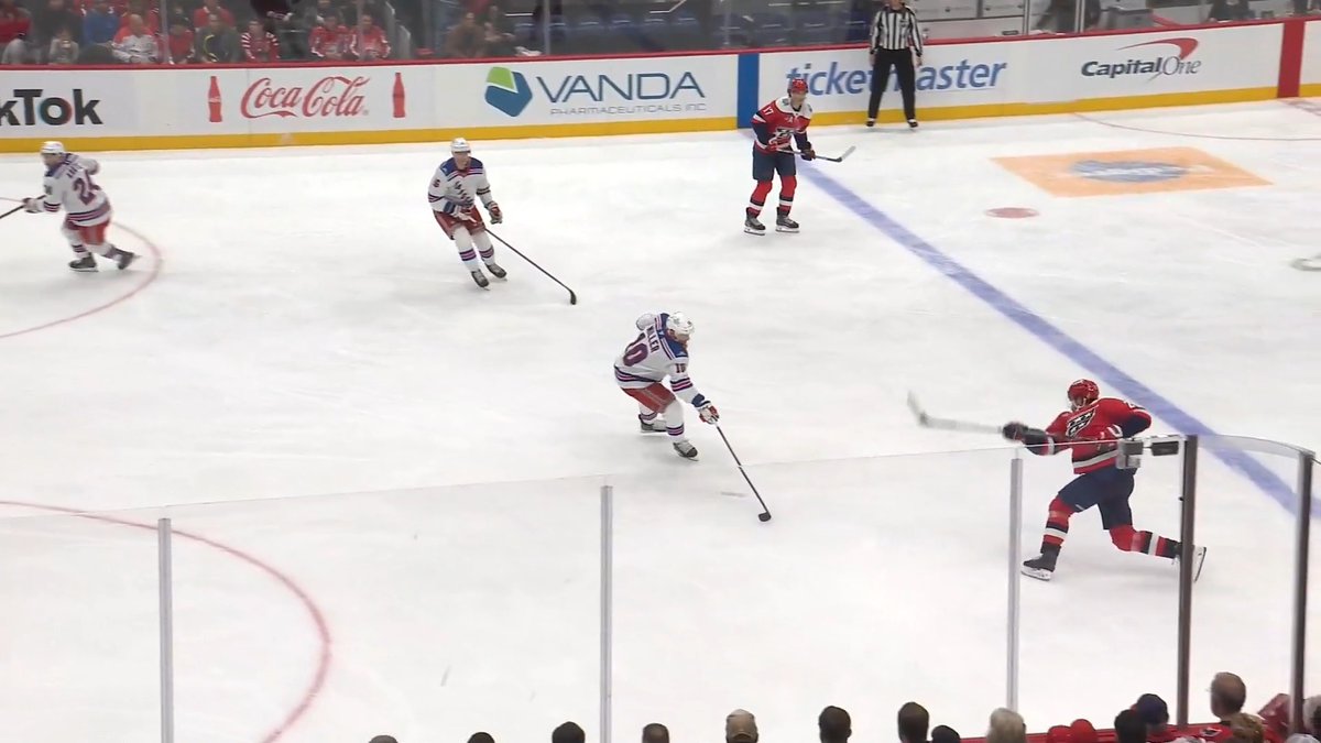 Hockey players from the Capitals (in red) and Rangers (in white) compete on the ice during a game, with the puck being shot toward the goal in a power-play situation. The image captures the moment of "Lily's first goal as a Cap is a laser," showing the decisive shot that resulted in the goal.
