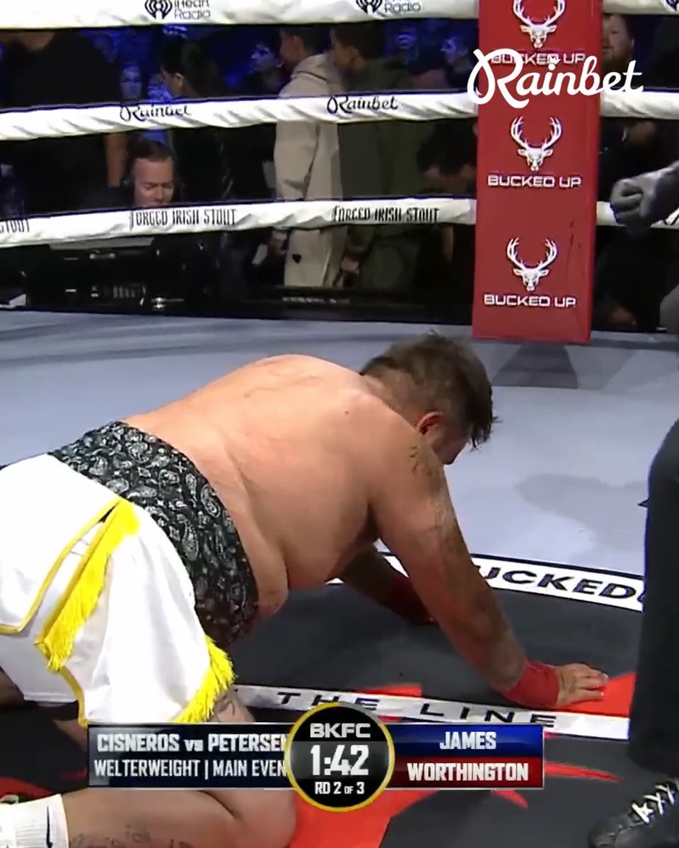 A bare-knuckle boxing match from BKFC (Bare Knuckle Fighting Championship) showing a fighter named Cisneros in white and yellow trunks on his knees during the main event against Petersen, with 1:42 remaining in Round 2 of 3, sponsored by Rainbet and Bucked Up.