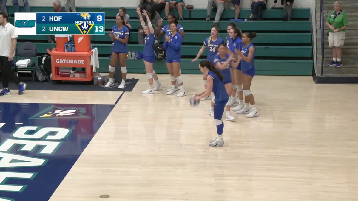 Hofstra volleyball team in blue uniforms celebrating a point during a match against UNCW, with the scoreboard showing HOF 2 (9) and UNCW 2 (13).