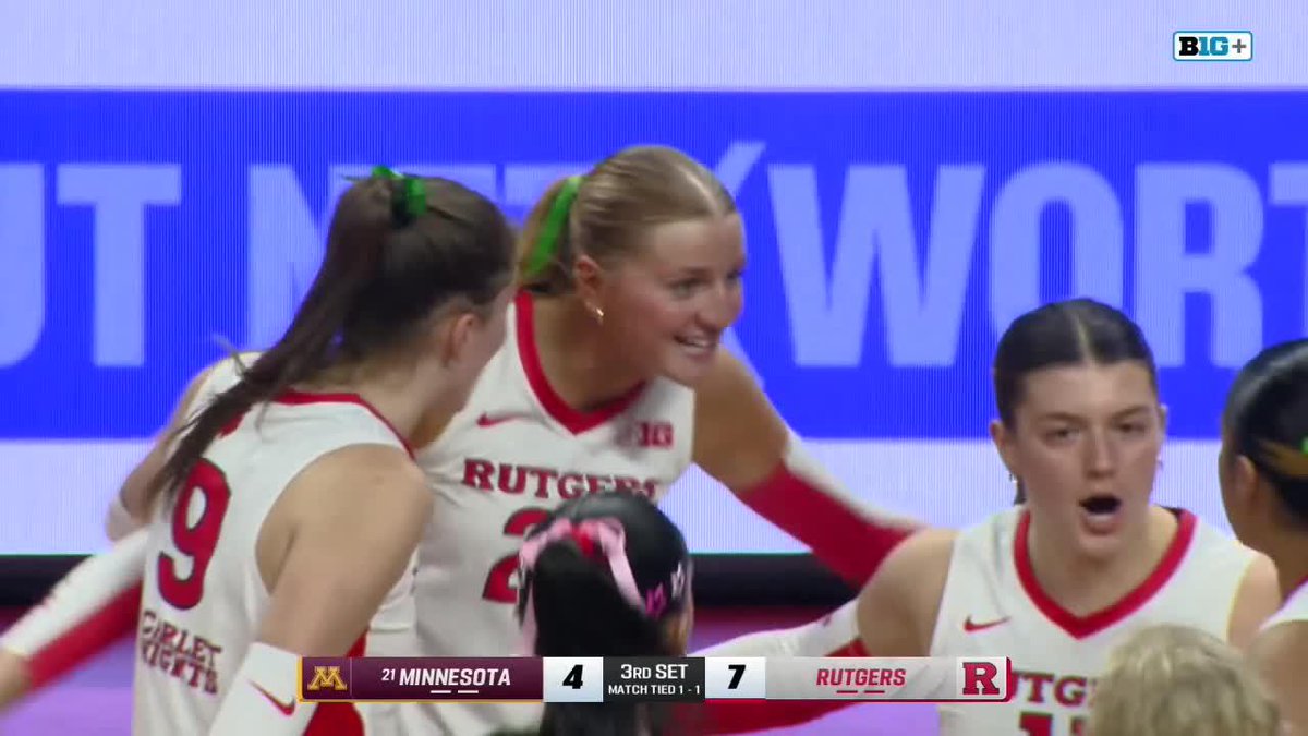Rutgers women's volleyball players in white and red uniforms celebrate during a match against #21 Minnesota, with Rutgers leading 7-4 in the 3rd set.