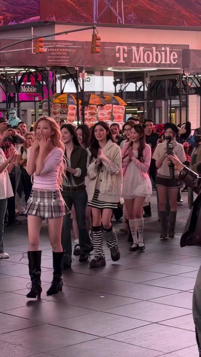 A group of young women, led by a woman with long strawberry-blonde hair in a pink top and plaid skirt, perform in what appears to be a public square with a T-Mobile store in the background, surrounded by a crowd of onlookers.