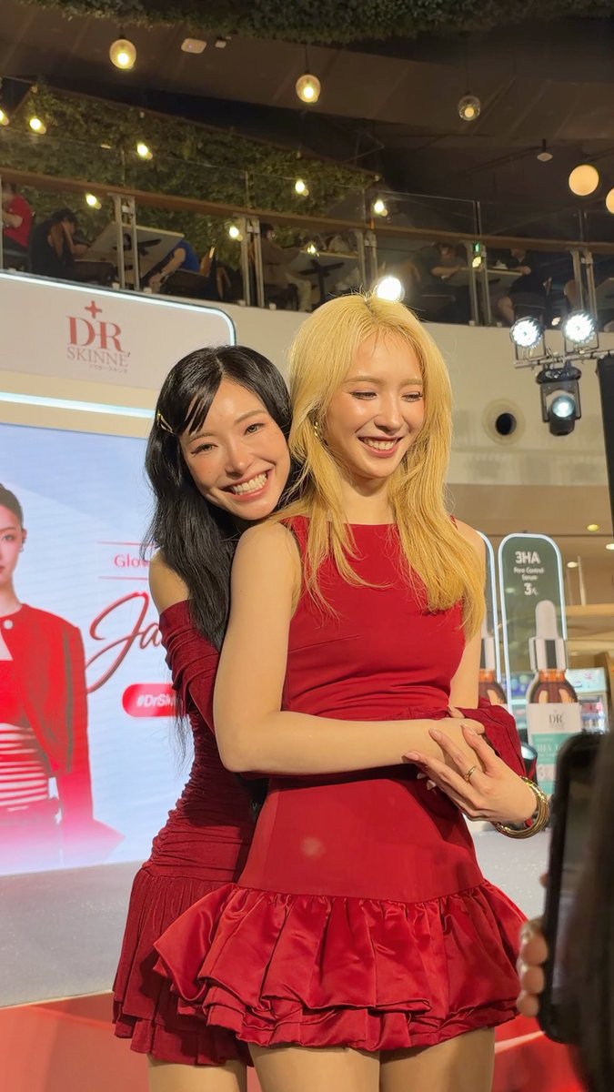 Two smiling women embracing at what appears to be a DR SKINNE promotional event; one has long black hair and the other has blonde hair, both wearing matching red dresses.