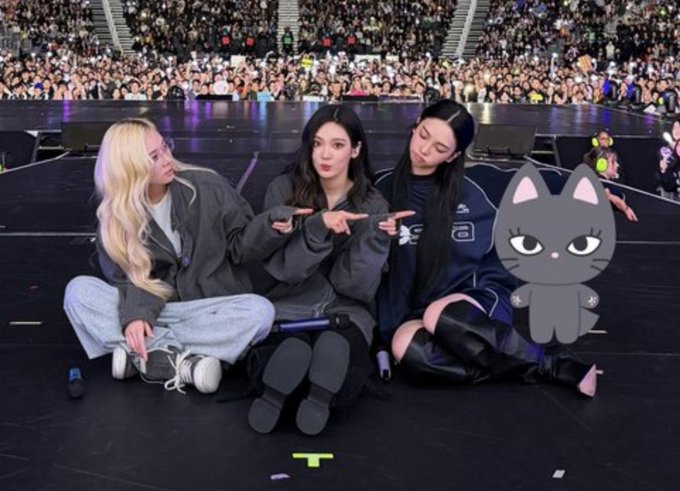 Three women sit on the floor of a stage in front of a large audience, posing together while a cartoon cat sticker covers part of the image.