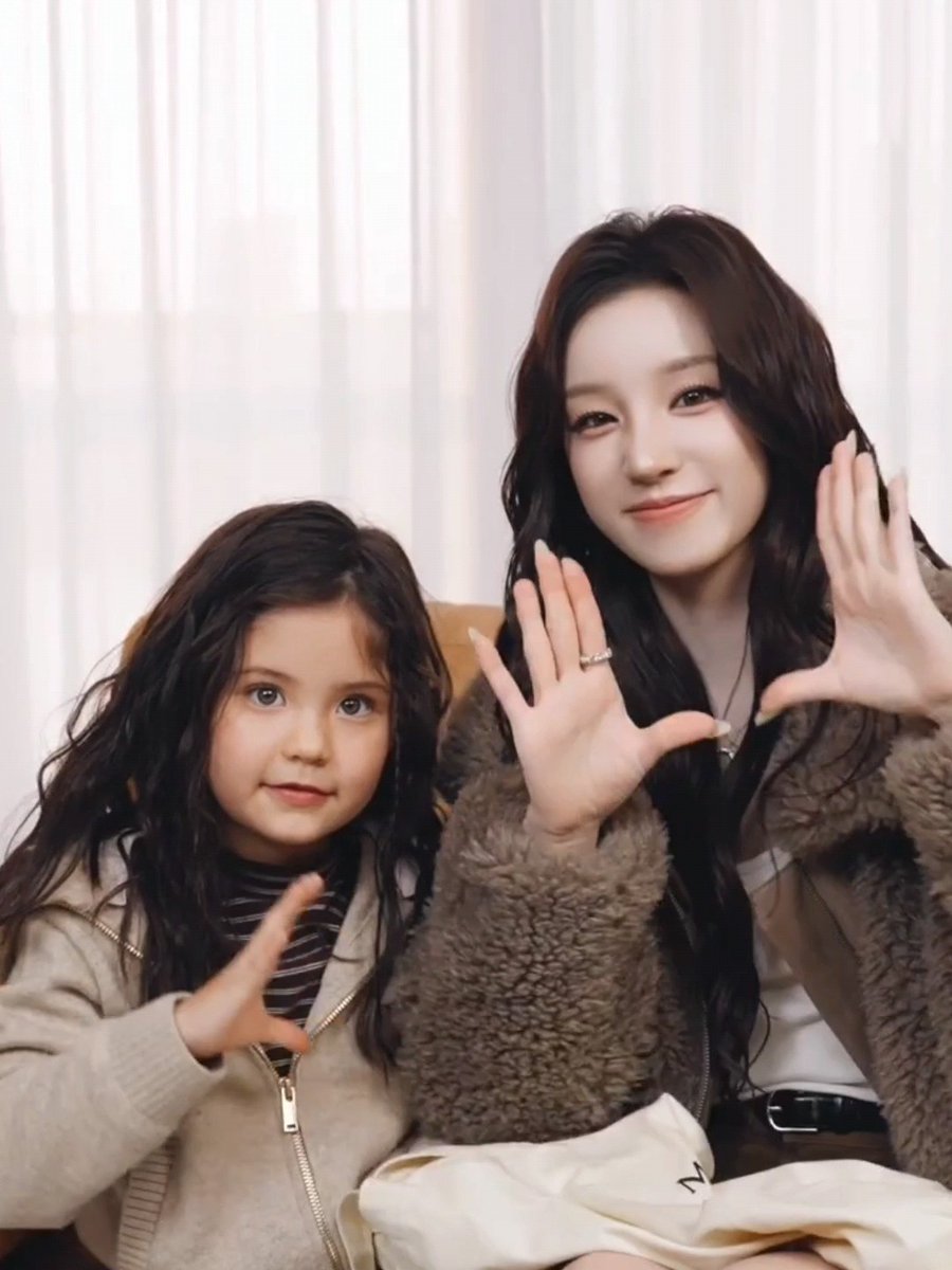 Two young women with long dark hair smile and wave at the camera; the older one on the right wears a fluffy brown jacket, and the younger one on the left wears a beige zip-up.