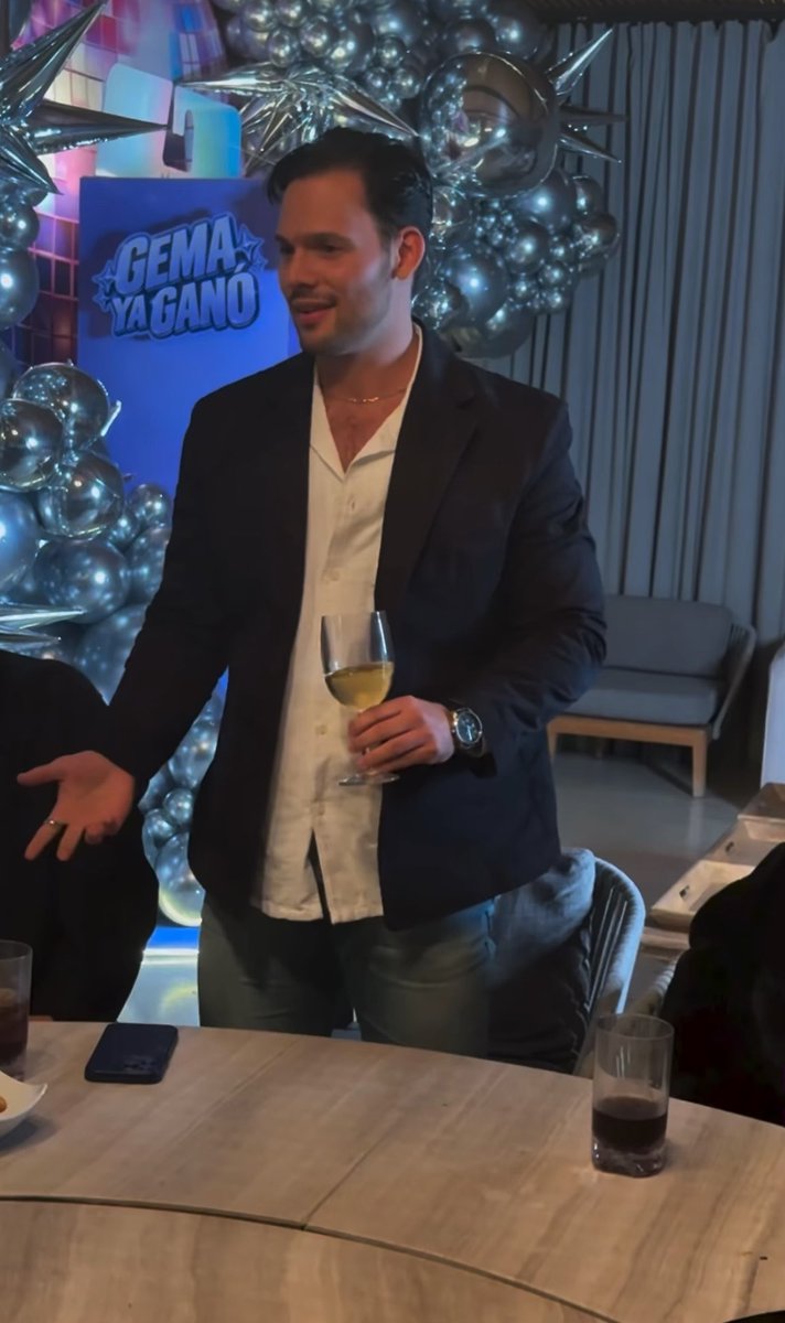 A smiling man in a dark blazer and white shirt holding a glass of white wine at an event, standing before silver balloons and a sign reading "GEMA YA GANO."