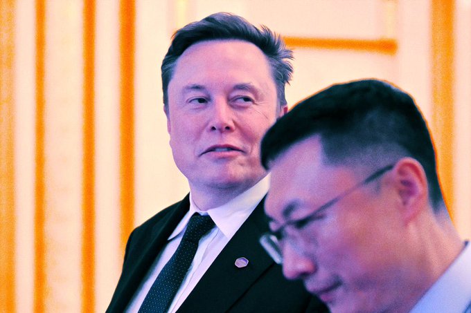 Elon Musk in a black suit and tie stands next to another man wearing glasses in an official setting.