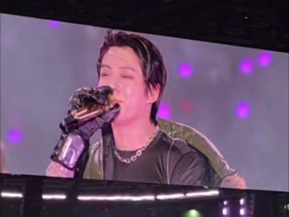 A young male performer with highlighted hair sings passionately into a microphone wearing a black glove and a black shirt with a silver chain, appearing on a stage screen.