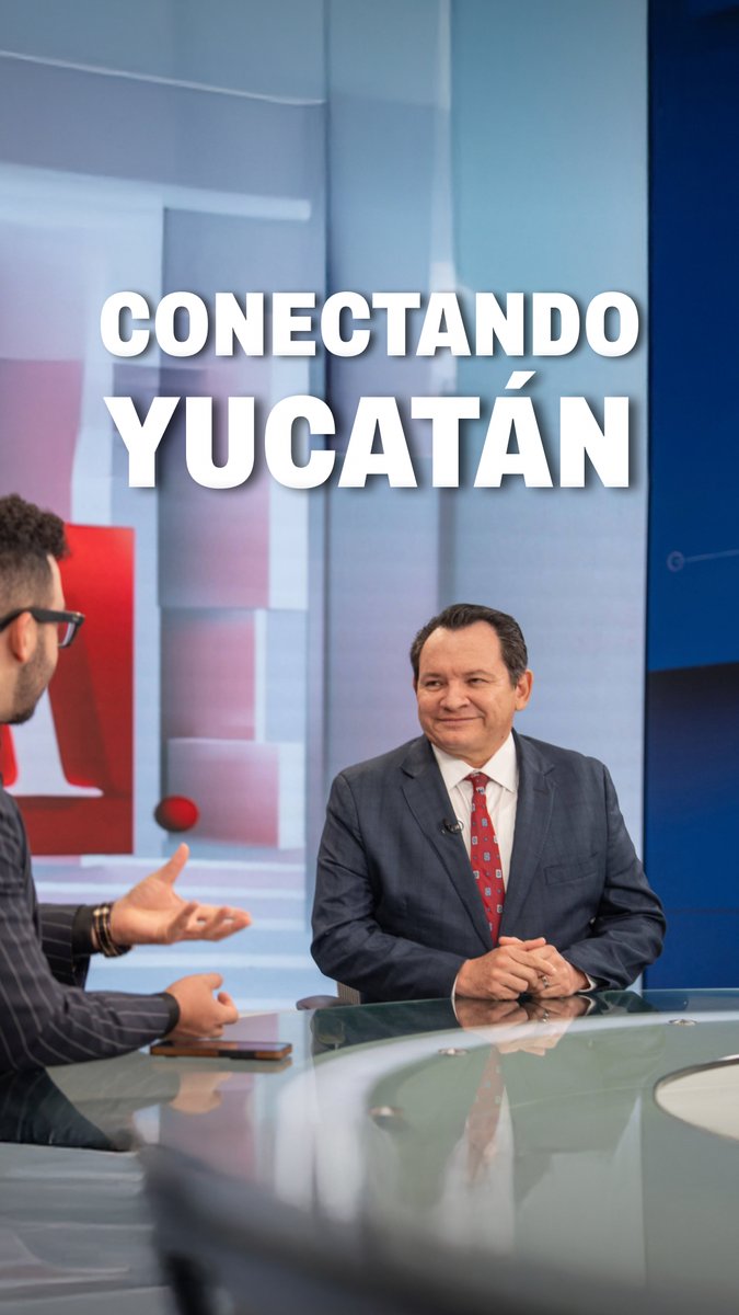 A television interview screenshot titled 'CONECTANDO YUCATÁN' showing two men at a glass studio desk, with one in a suit and red tie smiling while the other, wearing sunglasses and a striped jacket, gestures toward him.