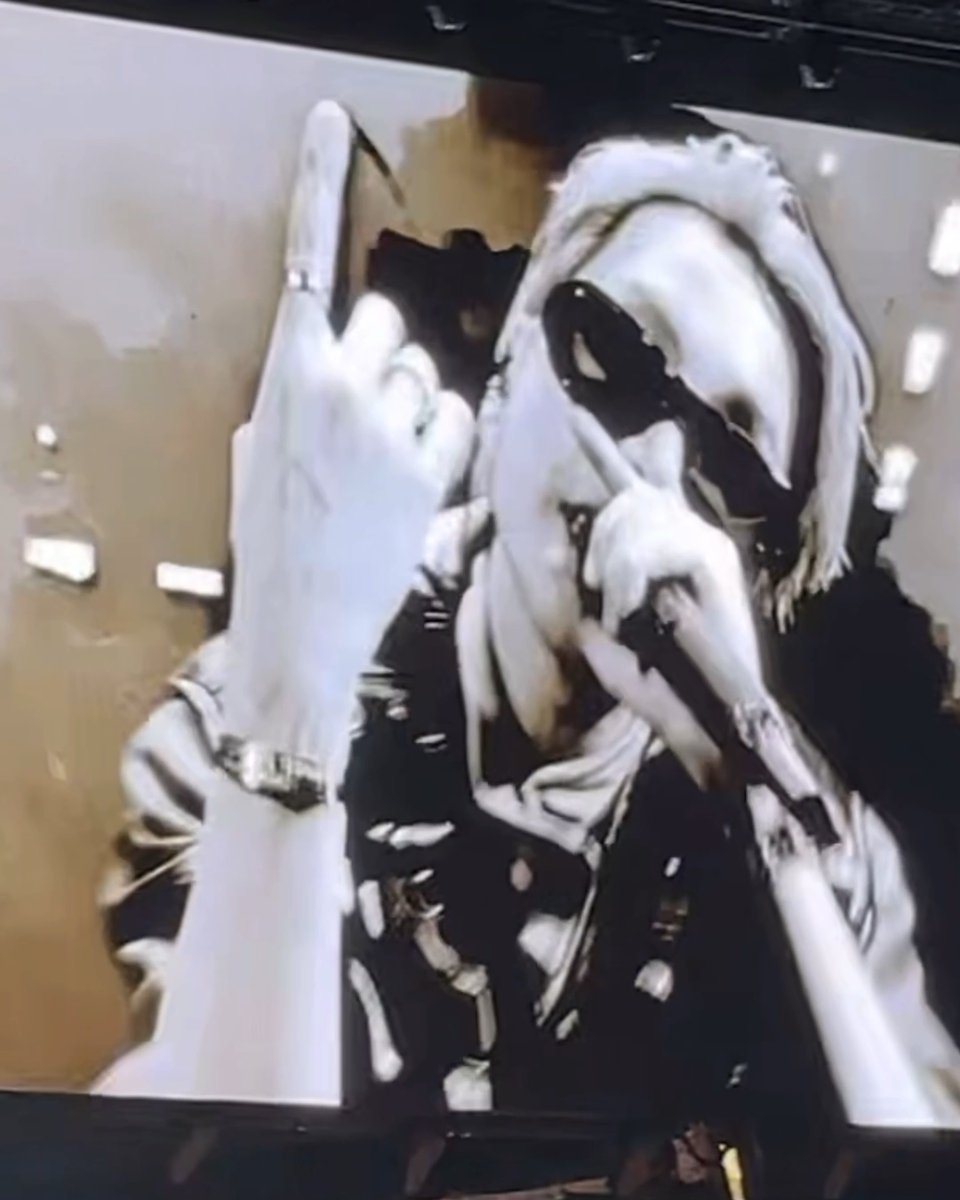 A distorted, glitchy image shows a figure in white with long blonde hair and sunglasses, appearing to hold a small object in their hand against an abstract background.