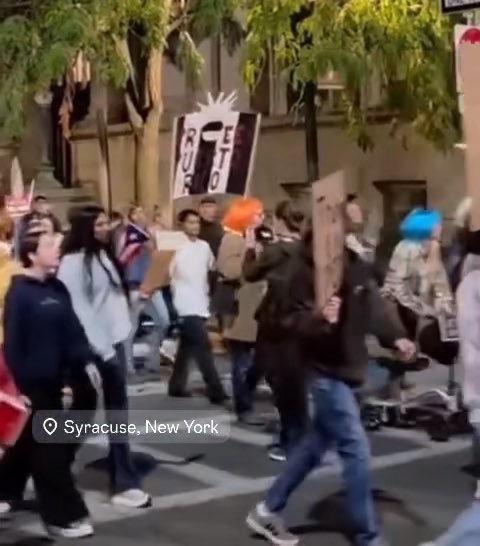 A photograph of a protest or parade in Syracuse, New York, with marchers carrying signs, including a visible 'OTF BO' sign, and a person wearing an orange wig walking through the crowd.