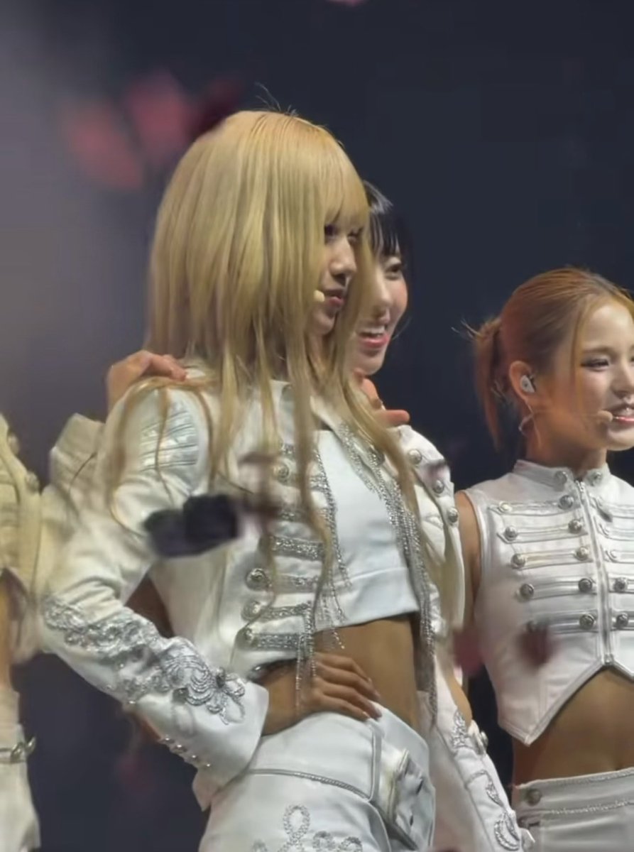 A group of female K-pop performers in white military-style stage outfits with silver embellishments during a concert performance.