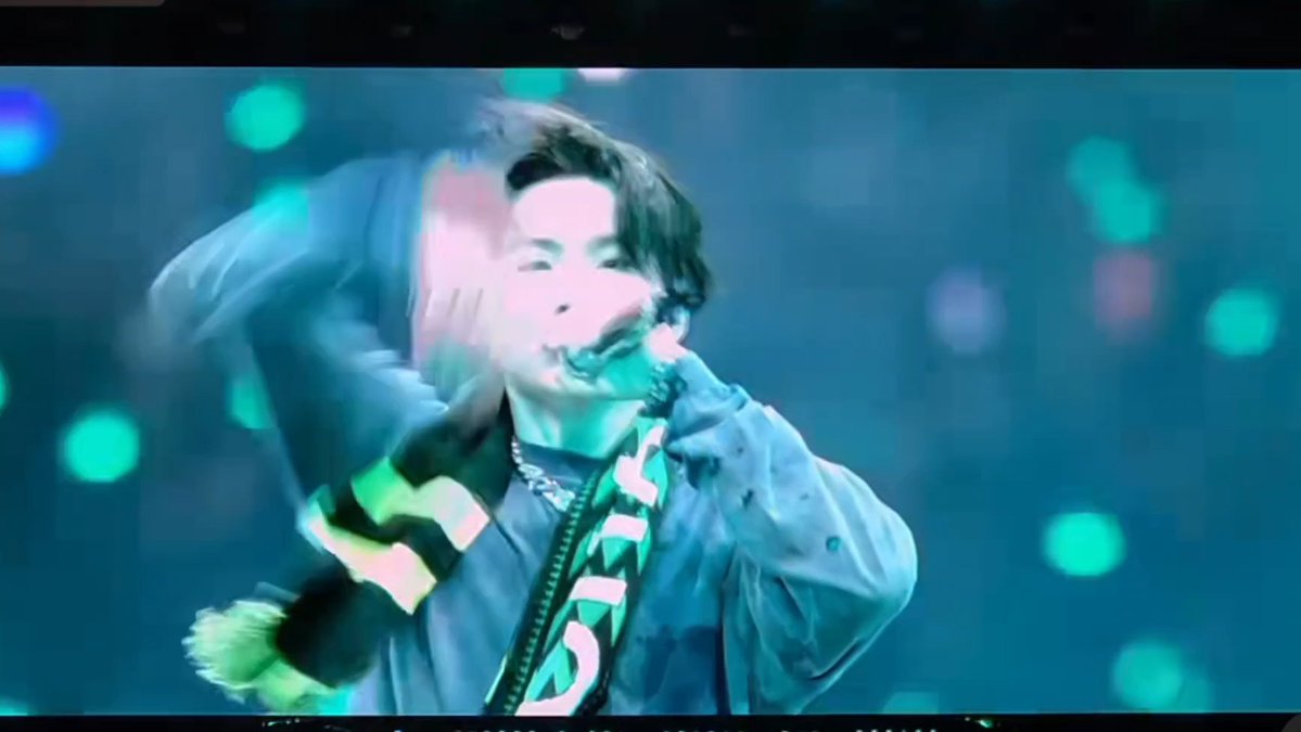 A blurry, stylized photo of a performer on stage singing into a microphone, wearing a light blue jacket with patterned clothing and a chain necklace, against a bokeh of green stage lights.