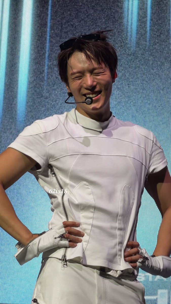A male performer with sunglasses perched on his head smiles broadly while wearing a futuristic white sleeveless outfit with zippers and fingerless gloves, with the watermark '62is18' visible.