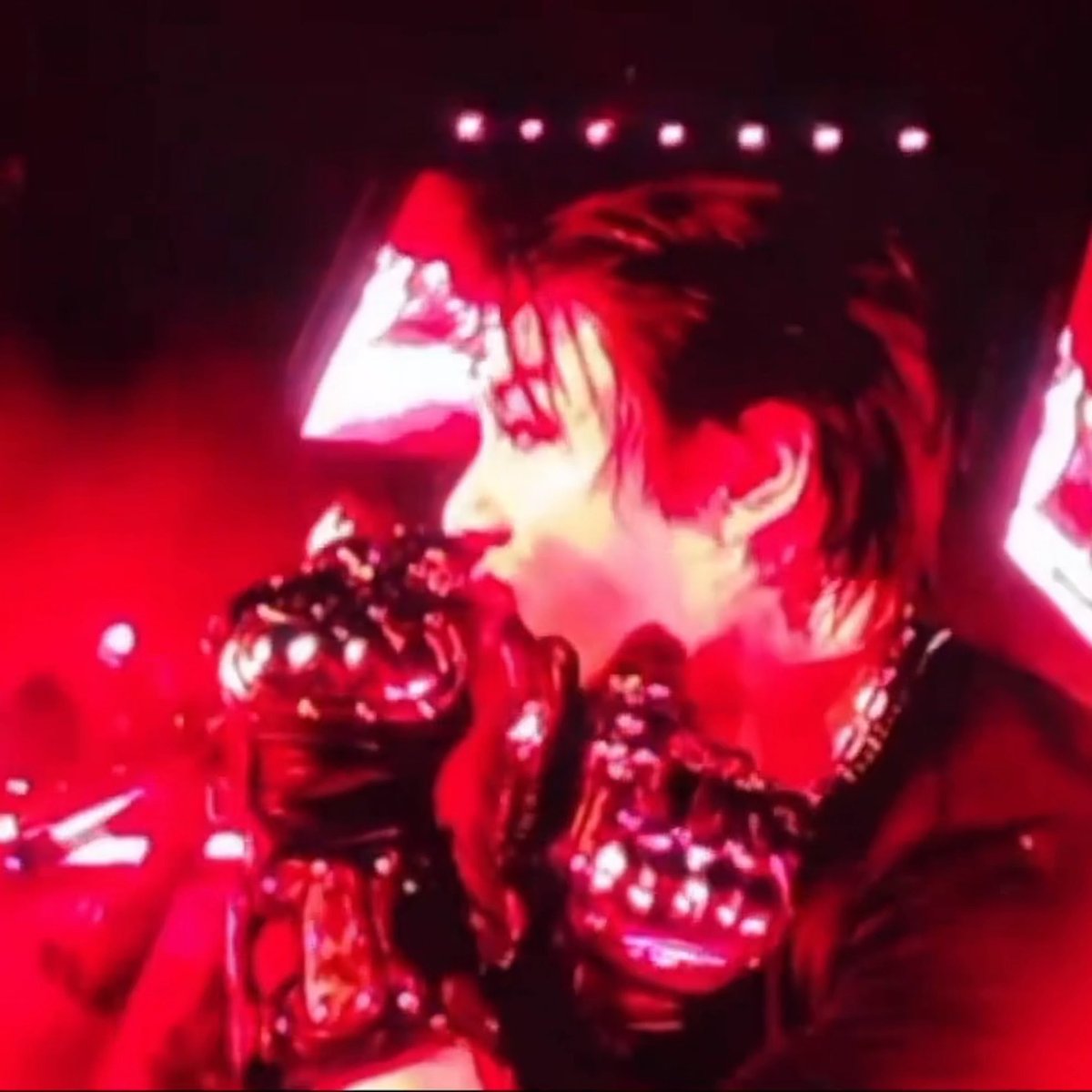 A blurry, red-tinted concert photo showing a performer with short hair on stage under bright red lighting.