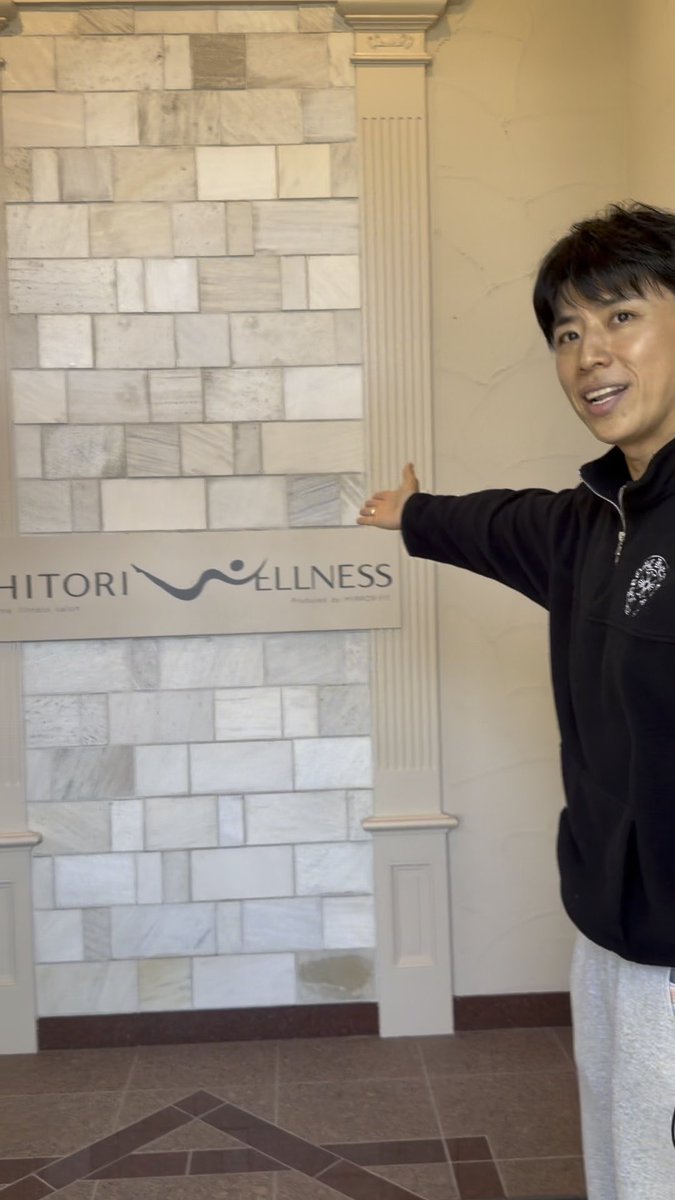 A man in a black zip-up jacket and gray sweatpants gestures toward a marble wall with a sign reading 'HITORI WELLNESS' and the tagline 'PHILOSOPHY FOR PERFECTION'.