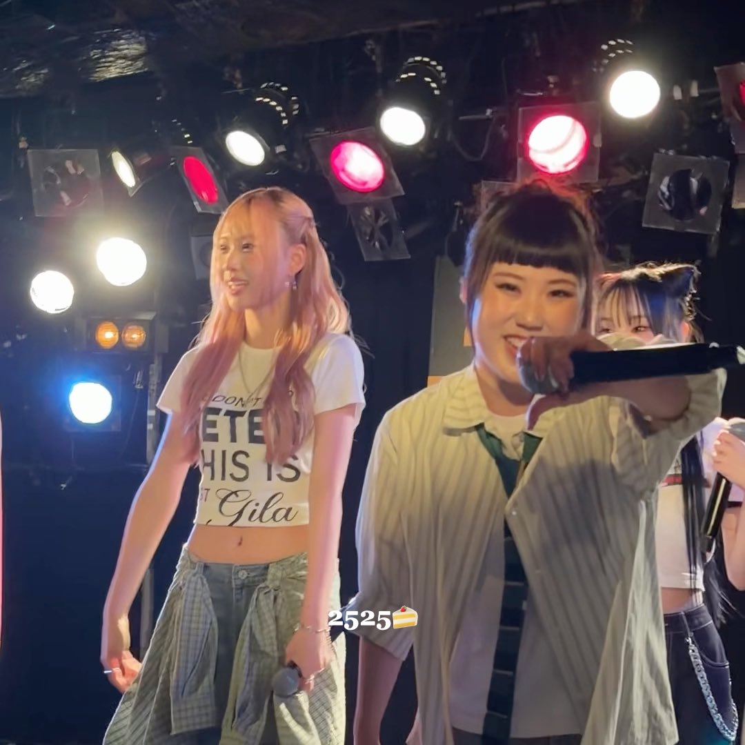 A concert performance featuring a blonde woman with pink-tipped hair in a white crop top and another performer holding a microphone on stage with colorful stage lights.