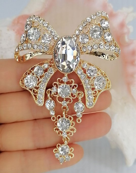 A hand holds an ornate gold-toned bow-shaped brooch featuring a large central oval crystal and intricate filigree decorated with numerous round clear stones.