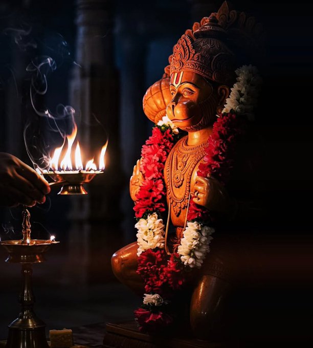 An ornate statue of the Hindu deity Hanuman adorned with red and white flower garlands, with a lit brass oil lamp held nearby in the foreground.