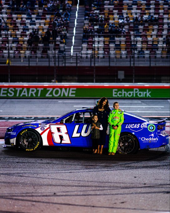 A NASCAR driver in a neon green suit stands alongside a woman and child in front of a blue #8 race car sponsored by Lucas Oil and Cheddar's, with "RESTART ZONE" and "EchoPark" signage visible in the background—capturing a family moment at the track that reflects the post's sentiment, "Always our family."