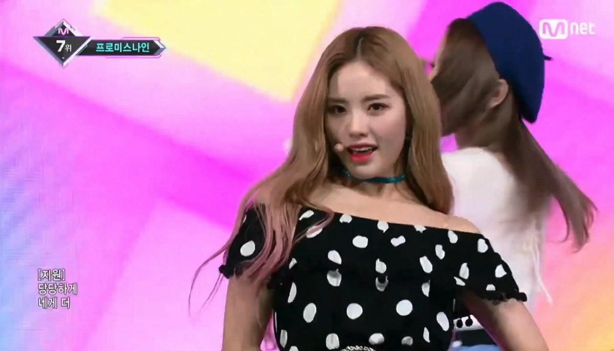 A screenshot from an Mnet broadcast showing a woman with light brown hair in a black polka dot off-shoulder top performing on stage, with a '프로미스나인' (fromis_9) logo and Korean subtitles visible.