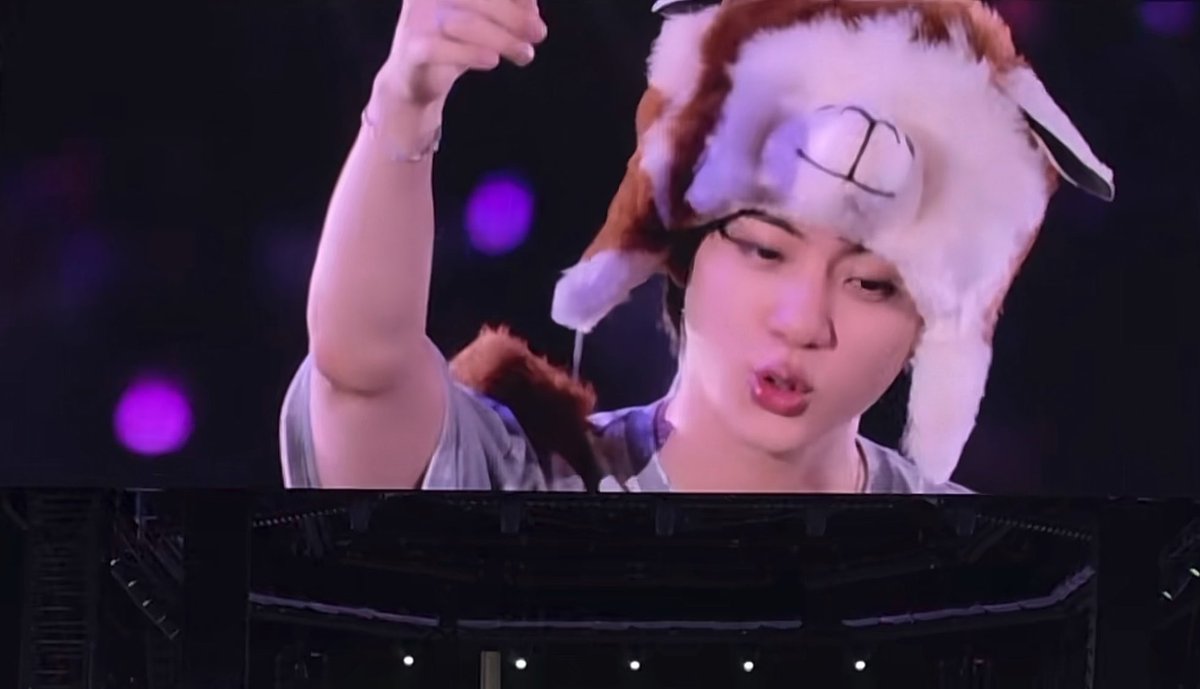 A performer wearing a furry animal ear hat is shown on a large stage screen, captured mid-motion with one arm raised.