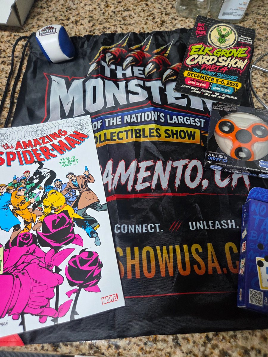 Various collectibles laid out on a granite countertop, including a 'The Monster' drawstring bag from the Sacramento Collectibles Show, an Amazing Spider-Man comic book, a promotional flyer for an 'Elk Grove Card Show', and an orange fidget toy.