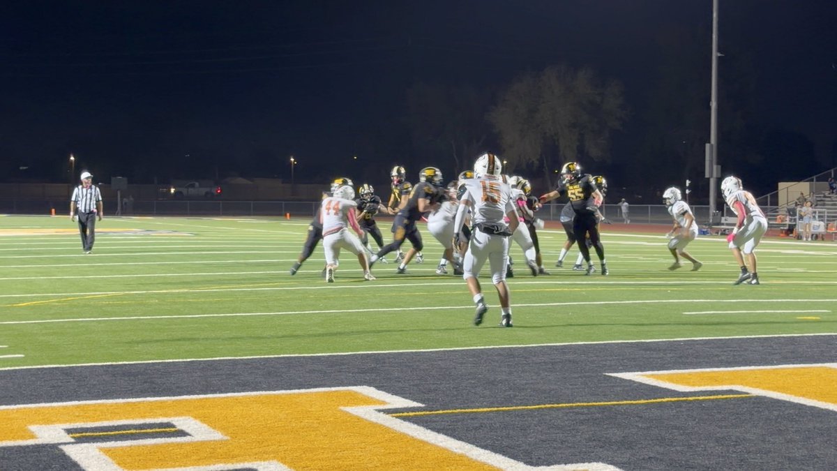 A nighttime high school football game in progress, with players in white and dark jerseys lined up at the line of scrimmage while a referee observes from the sideline.