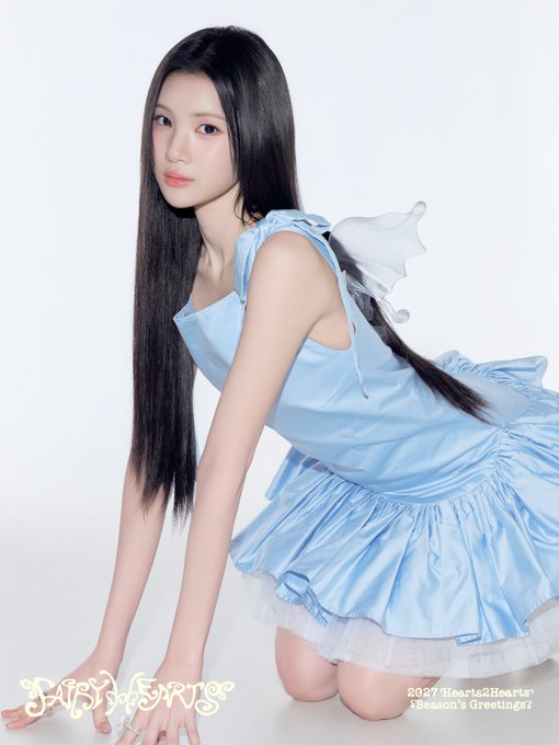 A promotional photo of a K-pop idol wearing a light blue dress with butterfly wings attached to the back. The image includes text for 'Hearts2Hearts' and reads '2025 Hearts2Hearts 1 Season's Greetings'.