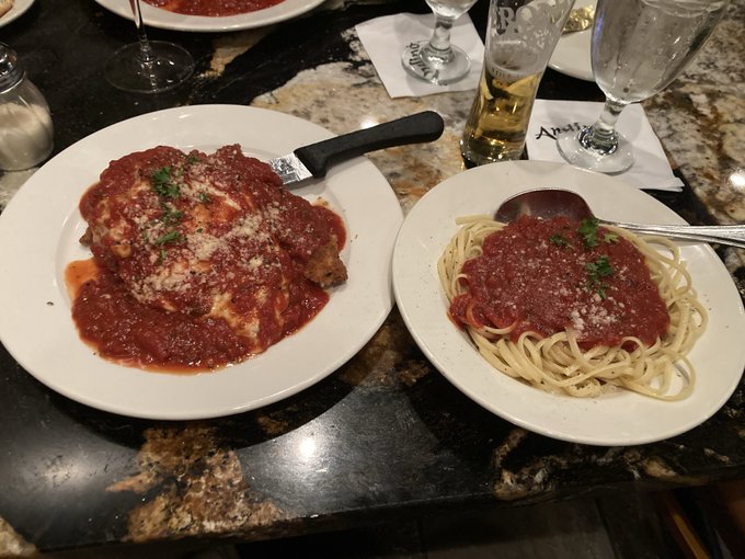 An Italian meal featuring chicken parmesan covered in marinara sauce and a plate of spaghetti with marinara sauce on a restaurant table with a knife and glasses.