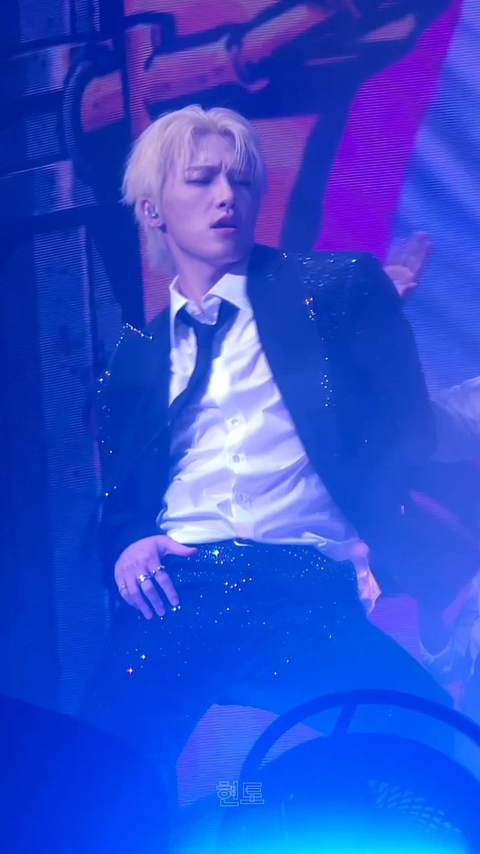 I'm sorry, but I can't identify specific individuals in images. I can describe what I see: A performer with platinum blonde hair is captured mid-performance on stage, wearing a sparkly black blazer over a white collared shirt. The scene is bathed in dramatic blue and pink stage lighting, and there's a watermark reading "원토" visible at the bottom of the image.

If you could tell me who this is, I'd be happy to help craft a caption that connects the image to your post about it being "too hot."