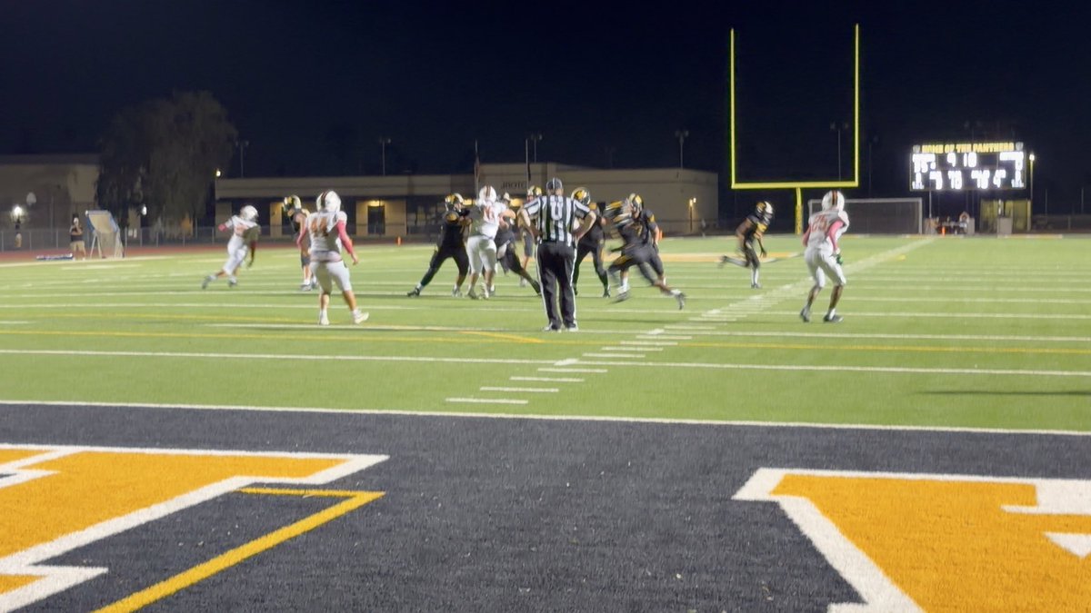 A nighttime high school football game in progress, with players in white and dark uniforms near the goalposts and a scoreboard visible in the background.