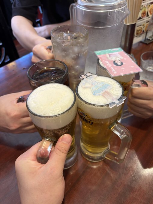 A first-person view of people toasting with beer mugs at a restaurant table, featuring two frothy mugs in the foreground clinked together, with other drinks and a menu visible on the wooden table.