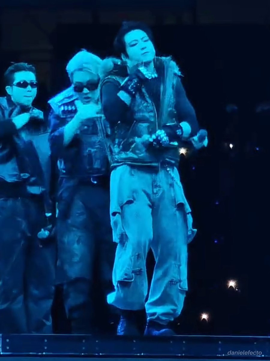 Concert photo showing performers in dark tactical-style outfits under blue stage lighting, watermarked 'danielefecto'.