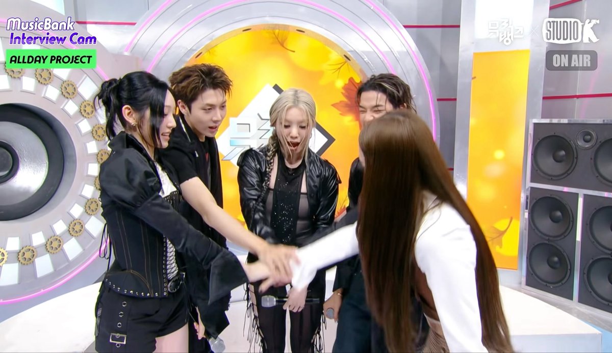 The image shows the K-pop group ALLDAY PROJECT on Music Bank's Interview Cam, with the text "MusicBank Interview Cam ALLDAY PROJECT" visible in the corner and a "STUDIO K ON AIR" sign displayed, matching the hashtagged post celebrating members Bailey, Tarzan, Woochan, Youngseo, and Annie as they share a playful moment with joined hands in the studio.