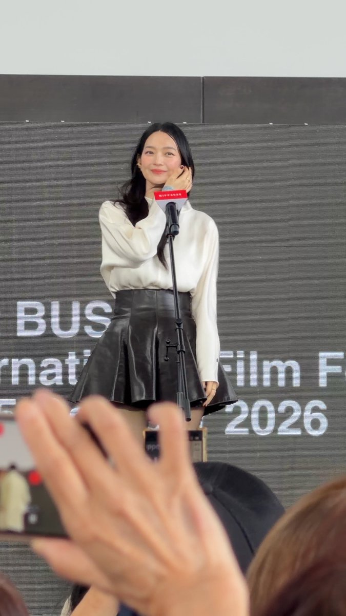 A woman in a white blouse and black leather skirt stands at a microphone at what appears to be the Busan International Film Festival 2026.