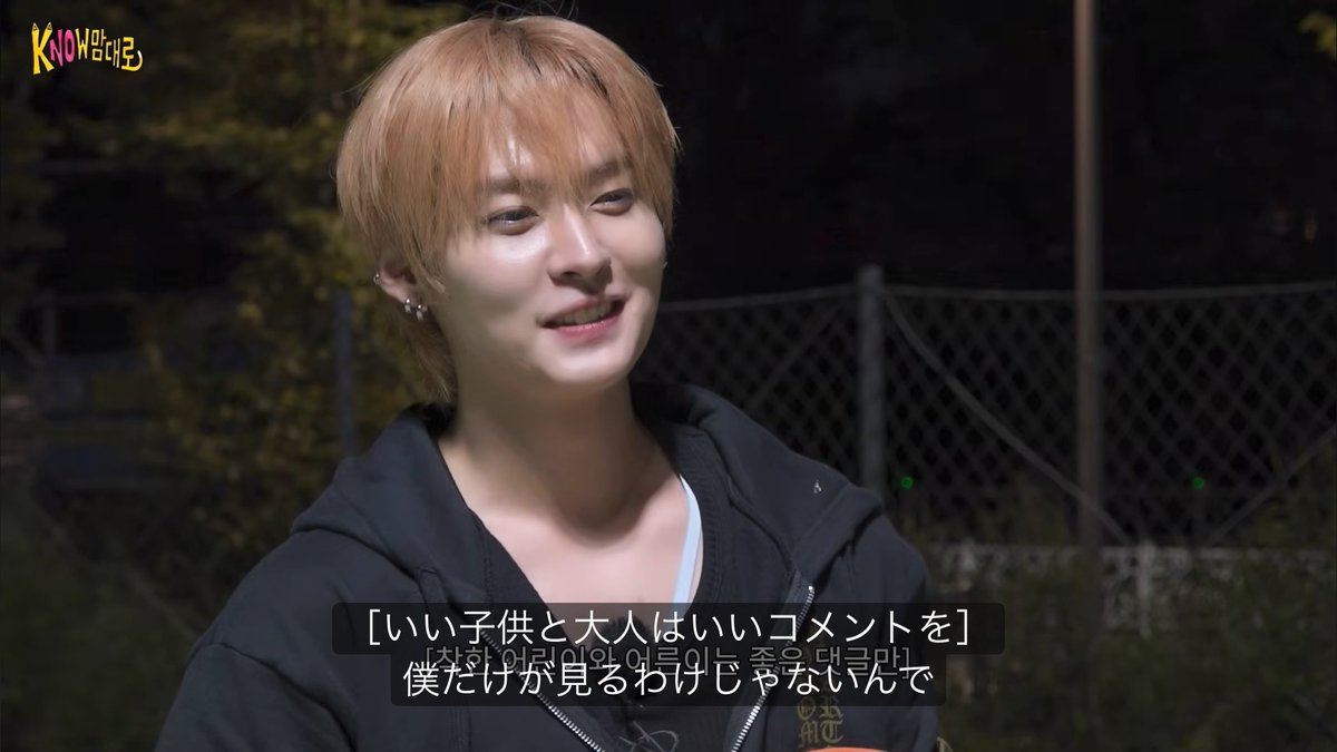A young man with orange hair, smiling softly during an outdoor nighttime interview with a "KNOW" logo in the corner, appears alongside Japanese subtitles reading "僕だけが見るわけじゃないんで" (meaning "It's not just me watching"), reflecting the accompanying message about leaving thoughtful comments for everyone to see, not just for himself.